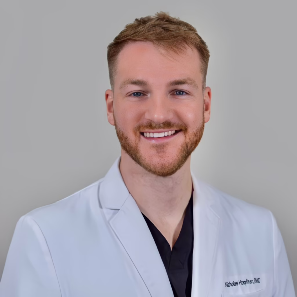Portrait of a smiling male doctor or clinician wearing a white coat, head and shoulders against a neutral background.