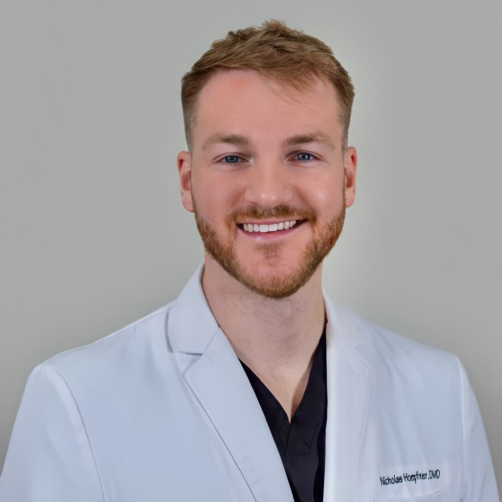 Portrait of a smiling male doctor or clinician wearing a white coat, head and shoulders against a neutral background.